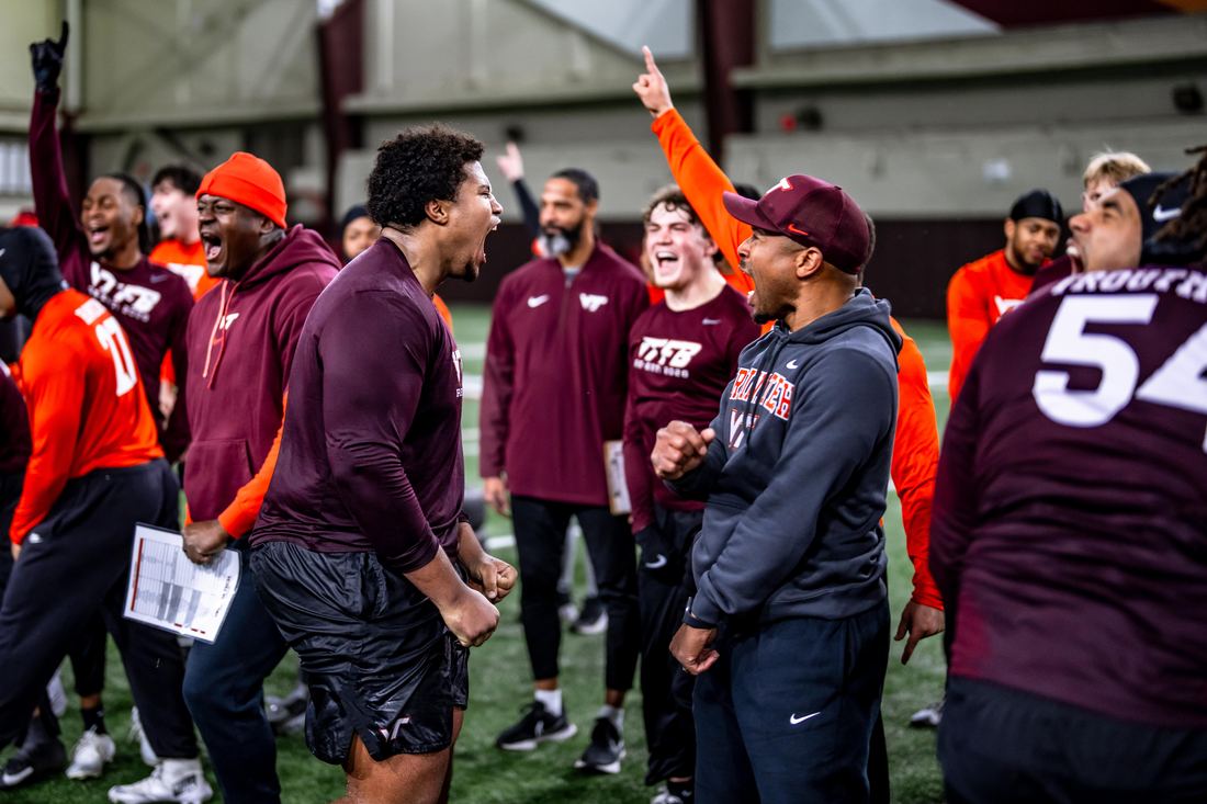 Benjamin Eziuka shouting in celebration face-to-face with a Virginia Tech coach, teammates cheering around them in the indoor facility.