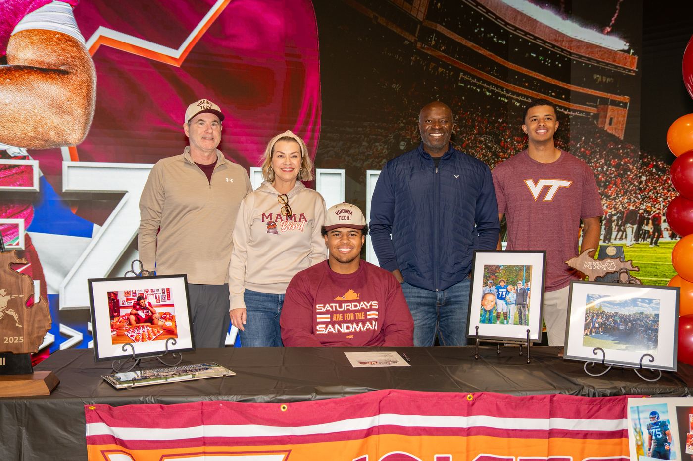Signing day: Benjamin Eziuka seated at a table in a Virginia Tech cap with his commitment paperwork, family standing behind him and the state championship trophy on the table.